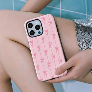 Op de afbeelding: Roze telefoonhoesje met een herhalend patroon van lichtroze strikken op een roze achtergrond. De hoes heeft een zwarte rand en een camera-uitsparing. De telefoonhoes wordt in de hand van een persoon gehouden.