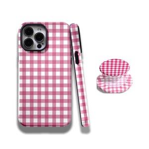 Pode incluir: Uma capa de telefone e um suporte a condizer com um padrão xadrez rosa e branco, e um telefone. A capa e o suporte apresentam um padrão xadrez clássico em estilo piquenique. A capa foi concebida para um smartphone.