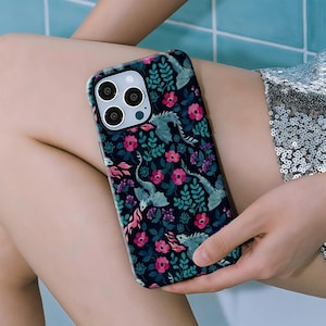May include: A phone case with a dark background featuring a floral and dragon pattern. The design includes pink flowers, green leaves, and light blue dragons. The case has a black border and camera cutouts.