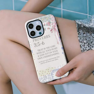 May include: A floral patterned phone case with the text "PROVERBS 3:5-6" and a Bible verse. The case features a pastel color scheme with pink and blue floral accents and a pink ribbon design. The phone case is held by a person.
