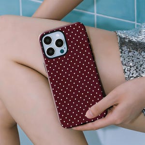 May include: A burgundy phone case featuring a white polka dot design. The case has a camera aperture and is held in a person's hand. The phone case is a solid colour with a repeating pattern of small white dots.