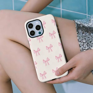 Op de afbeelding: Een crèmekleurige telefoonhoes met een patroon van roze strikken. De hoes heeft een zwarte rand en een uitsparing voor de camera. De strikken zijn gelijkmatig verdeeld over het oppervlak van de hoes.