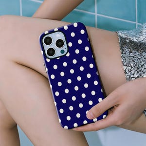 Op de afbeelding: Een marineblauwe telefoonhoes met een wit stippenpatroon. De hoes heeft een camera-uitsparing en wordt in een hand gehouden. Deze telefoonhoes is een beschermend accessoire voor een smartphone.