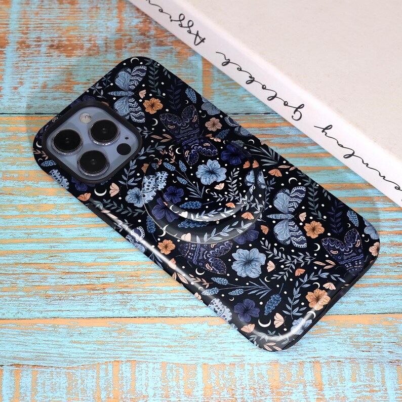 May include: Black phone case with a floral and butterfly pattern in shades of blue, orange, and white. The case has a built-in phone grip. The phone case is lying on a light blue and brown wooden surface.