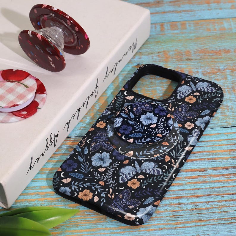 May include: A black phone case with a floral and butterfly pattern, featuring a matching pop-up grip. The pop-up grip and a separate round grip have a red floral design. The items are on a light blue and wood-grain surface.