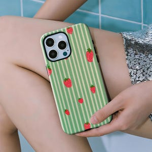 May include: A green and white striped phone case featuring a pattern of red strawberries. The case has a camera cutout and is designed to fit a smartphone. The overall design is a playful and summery theme.