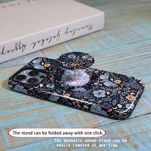 May include: A black phone case with a floral and butterfly pattern, featuring a matching pop-up phone grip. The grip is clear with a floral design. The text on the image says, "The stand can be folded away with one click."