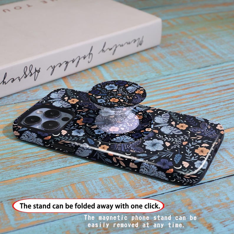 May include: A black phone case with a floral and butterfly pattern, featuring a matching pop-up phone grip. The grip is clear with a floral design. The text on the image says, "The stand can be folded away with one click."
