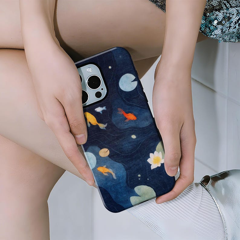 May include: A dark blue phone case featuring a pond scene with orange and yellow koi fish, lily pads, and a white lotus flower. The case has a glossy finish and is held by a person.