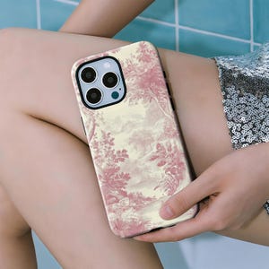May include: A phone case with a cream-colored background and a pink toile pattern featuring trees and landscapes. The case has a black border and a camera cutout. The phone case is held in a person's hand.