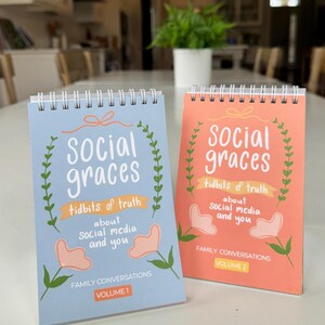 May include: Two spiral-bound notebooks with the text "Social Graces" and "tidbits of truth about social media and you." Volume 1 is light blue, and Volume 2 is coral. Both have floral illustrations and the words "Family Conversations."