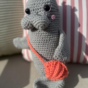 Puede incluir: Un manatí gris tejido a mano con grandes ojos negros, una pequeña mejilla rosa y una corona morada. Tiene un bolso de color coral y una base gris. Un artículo artesanal caprichoso.