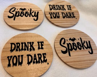 Op de afbeelding: Vier ronde houten onderzetters met zwarte tekst. Twee onderzetters lezen "Spooky" en de andere twee "Drink if You Dare". De onderzetters zijn gemaakt van lichtgekleurd hout met zichtbare nerf.