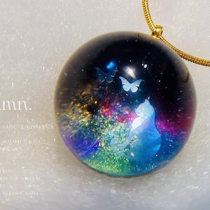 Puede incluir: Colgante redondo con cadena dorada. El colgante transparente presenta un fondo azul oscuro y morado, una silueta de gato y dos diseños de mariposas. La palabra "Autumn" es visible en el fondo.