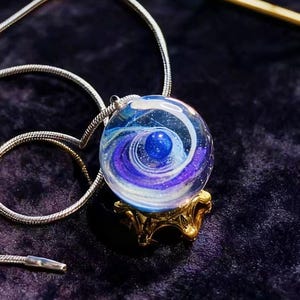 May include: A glass pendant necklace with a swirling blue and purple design, resting on a small gold-colored stand. The pendant is round with a small blue sphere in the center. The necklace chain is silver.