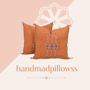 Può includere: Due cuscini quadrati arancioni con un motivo geometrico centrale ricamato in bianco, blu e rosa, con piccole nappe agli angoli. Il testo handmadepillowss è visualizzato sotto i cuscini con un carattere di colore arancione chiaro.
