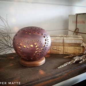 May include: A copper matte ceramic lamp with a spherical shape and a pattern of small holes. The lamp is lit and casting a warm glow. The lamp is sitting on a wooden base and is on a wooden shelf with books and dried flowers.