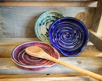 May include: Three ceramic spoon rests in different colors: a green and white swirl, a blue swirl, and a red and white swirl. A wooden spoon is resting on the red and white spoon rest.