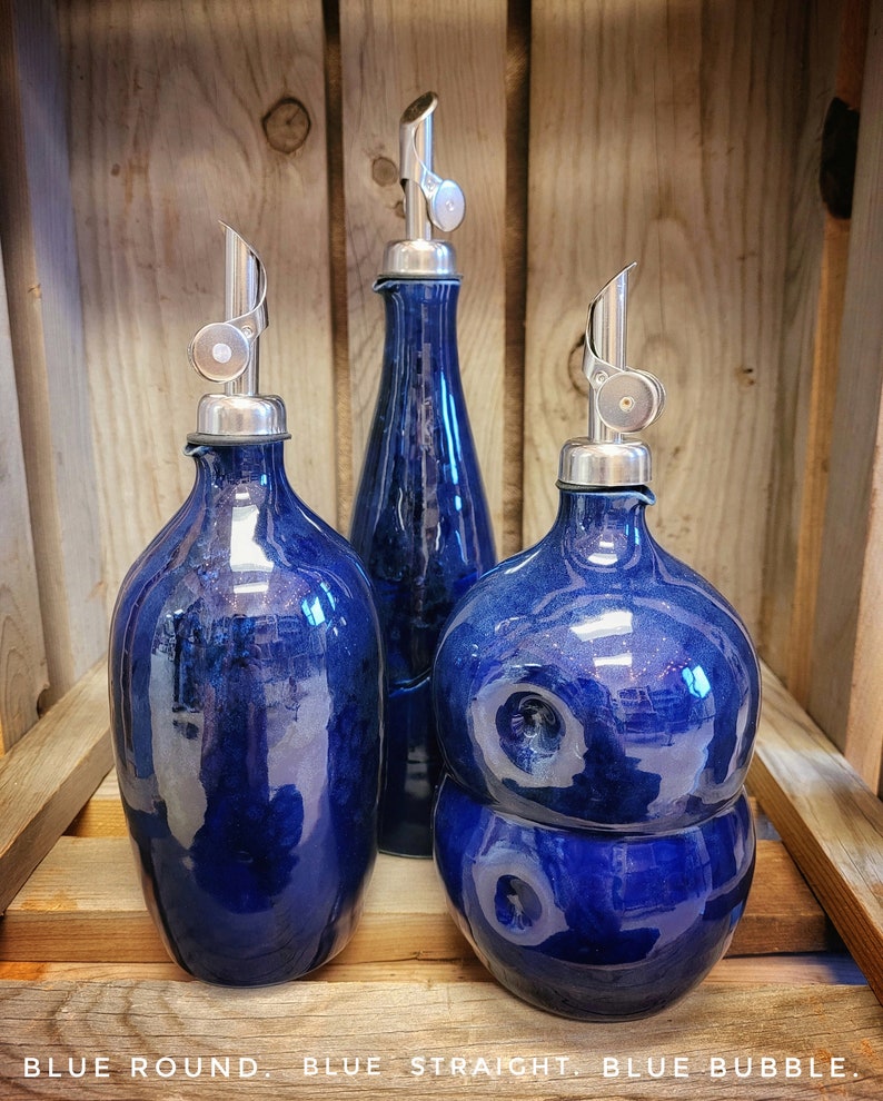 May include: Three blue ceramic oil and vinegar bottles with silver pour spouts. The bottles are in a wooden crate. The bottles are labeled "Blue Round", "Blue Straight", and "Blue Bubble".