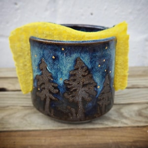 Puede incluir: Un soporte para esponja de cerámica con un esmalte azul y marrón, que presenta una escena de bosque con árboles en relieve y detalles dorados. Una esponja amarilla está colocada dentro. El soporte está sobre una superficie de madera.