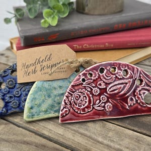 May include: Three semi-circular ceramic herb strippers in blue, light green, and deep red, each featuring embossed floral patterns and circular holes. A brown paper tag attached with twine reads Handheld Herb Stripper LING POTTERY.