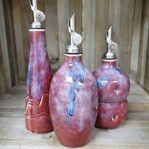 May include: Three ceramic oil and vinegar bottles with silver pour spouts. The bottles are a deep burgundy color with streaks of white and blue glaze. The bottles are arranged on a wooden surface.