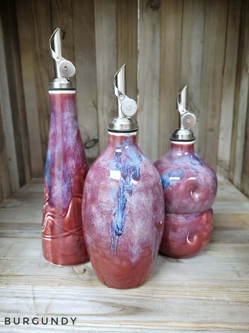 May include: Three ceramic oil and vinegar bottles with silver pour spouts. The bottles are a deep burgundy color with streaks of white and blue glaze. The bottles are arranged on a wooden surface.