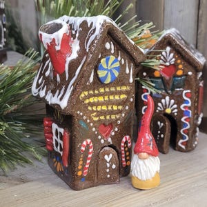 May include: Handmade ceramic gingerbread house with white icing, red chimney, and colorful candy decorations. A small gnome with a red hat and white beard stands in front of the house. The house is brown with yellow, blue, and red accents.