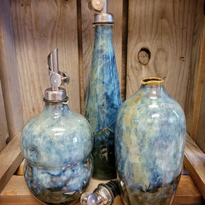 May include: Three ceramic oil and vinegar bottles with silver pour spouts. The bottles are in a blue and green glaze with a mottled effect. The bottles are labeled "Peacock Bubble", "Peacock Straight", and "Peacock Round".