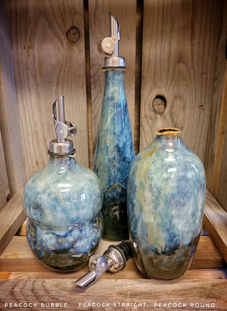 May include: Three ceramic oil and vinegar bottles with silver pour spouts. The bottles are in a blue and green glaze with a mottled effect. The bottles are labeled "Peacock Bubble", "Peacock Straight", and "Peacock Round".