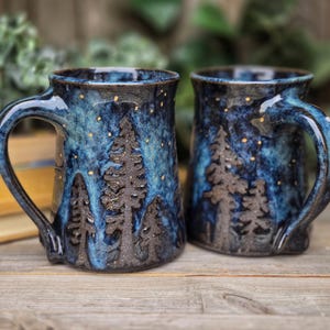 May include: Two blue and brown ceramic mugs featuring a forest scene with gold flecks. The mugs have handles and are ideal for hot drinks.