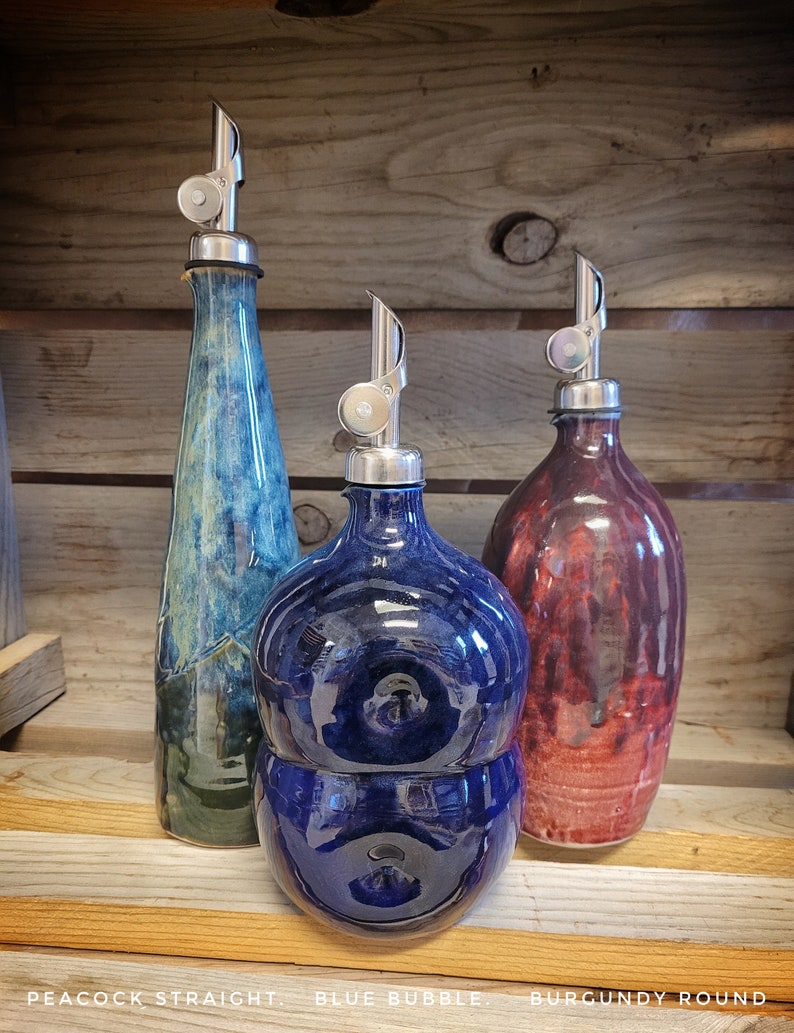 May include: Three ceramic oil and vinegar bottles with silver pour spouts. The bottles are in a peacock blue, a deep blue, and a burgundy color. The bottles are labeled "Peacock Straight", "Blue Bubble", and "Burgundy Round".