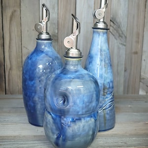 May include: Three blue ceramic oil and vinegar bottles with silver pour spouts. The bottles have a mottled blue glaze with white accents. The bottles are arranged on a wooden surface.