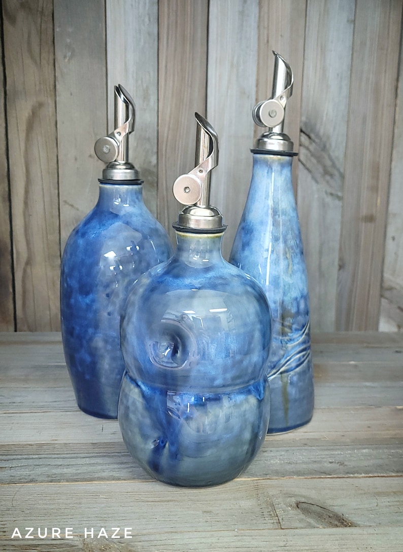 May include: Three blue ceramic oil and vinegar bottles with silver pour spouts. The bottles have a mottled blue glaze with white accents. The bottles are arranged on a wooden surface.