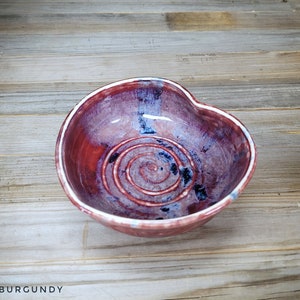 May include: A heart-shaped ceramic bowl with a white rim and a swirling pattern of burgundy, blue, and white glaze.