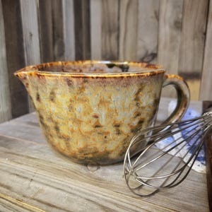 May include: A ceramic mixing bowl with a handle and spout, featuring a mottled brown and tan glaze. A stainless steel whisk rests beside the bowl. The bowl is designed for kitchen use.