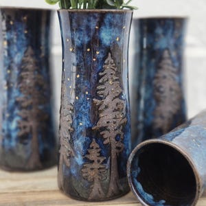 Op de afbeelding: Drie blauwe en bruine keramische vazen met een bos scène en gouden spikkels. De vazen staan op een houten oppervlak.