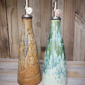 May include: Two ceramic oil and vinegar bottles with stainless steel pour spouts. The bottle on the left is brown with white and black marbling. The bottle on the right is green with white and blue marbling. The bottles are sitting on a wooden surface. "BROWN." and "GREEN." are written below the bottles.