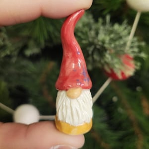 Puede incluir: Un pequeño adorno de gnomo de cerámica con un gorro rojo con detalles de copo de nieve azul, barba blanca y base amarilla.