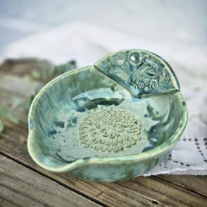 May include: A handcrafted, green ceramic dish with a floral design in the center. The dish has a unique shape with a raised, decorative section. The word "GREEN" is printed at the bottom of the image.