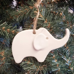 May include: White ceramic elephant ornament hanging from a brown twine string on a green artificial Christmas tree.