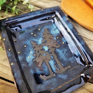 May include: A handmade, square ceramic dish with a dark blue glaze and a forest scene. The dish features two stylized trees in the center and small gold dots scattered across the surface. The edges are dark and the dish is on a wooden surface.