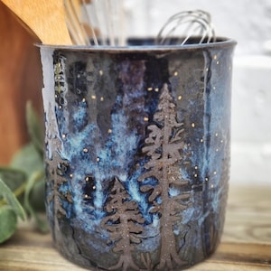 Puede incluir: Un porta utensilios de cerámica azul oscuro con un diseño de escena de bosque. El porta utensilios presenta un cielo nocturno estrellado con acentos de purpurina dorada y siluetas de pinos.