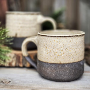 May include: Two ceramic mugs with a speckled beige upper half and a dark brown lower half. The mugs have a rounded handle and a dark brown rim. The background is blurred, with wood and greenery visible.