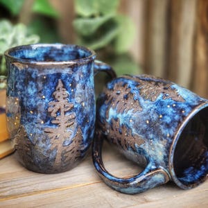 Puede incluir: Dos tazas de cerámica azul y marrón con un diseño de cielo estrellado y árboles. Las tazas tienen un esmalte moteado y un borde blanco.