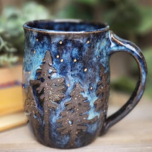 Puede incluir: Una taza de cerámica hecha a mano con un esmalte azul oscuro y negro. La taza presenta un diseño de pinos con un fondo de cielo estrellado.