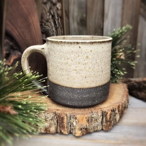 May include: A white ceramic mug with a speckled glaze and a black band around the top. The mug is sitting on a wooden slice.
