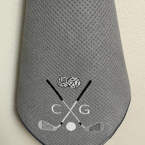 K&ouml;nnte beinhalten: Graues Golfhandtuch mit gesticktem Design. Das Design umfasst gekreuzte Golfschl&auml;ger, einen Golfball, W&uuml;rfel und die Buchstaben "C" und "G". Das Handtuch hat ein strukturiertes Muster und einen dunkelblauen Rand.