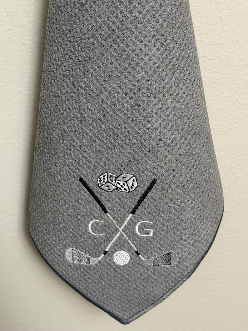 K&ouml;nnte beinhalten: Graues Golfhandtuch mit gesticktem Design. Das Design umfasst gekreuzte Golfschl&auml;ger, einen Golfball, W&uuml;rfel und die Buchstaben "C" und "G". Das Handtuch hat ein strukturiertes Muster und einen dunkelblauen Rand.