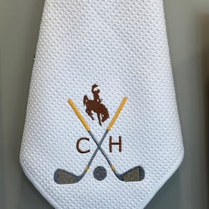 K&ouml;nnte beinhalten: Wei&szlig;es Golfhandtuch mit Waffelstruktur. Das Handtuch hat ein gesticktes Design mit gekreuzten Golfschl&auml;gern, einem Golfball und einer Cowboy-Silhouette auf einem Pferd. Die Initialen "C H" sind ebenfalls gestickt.
